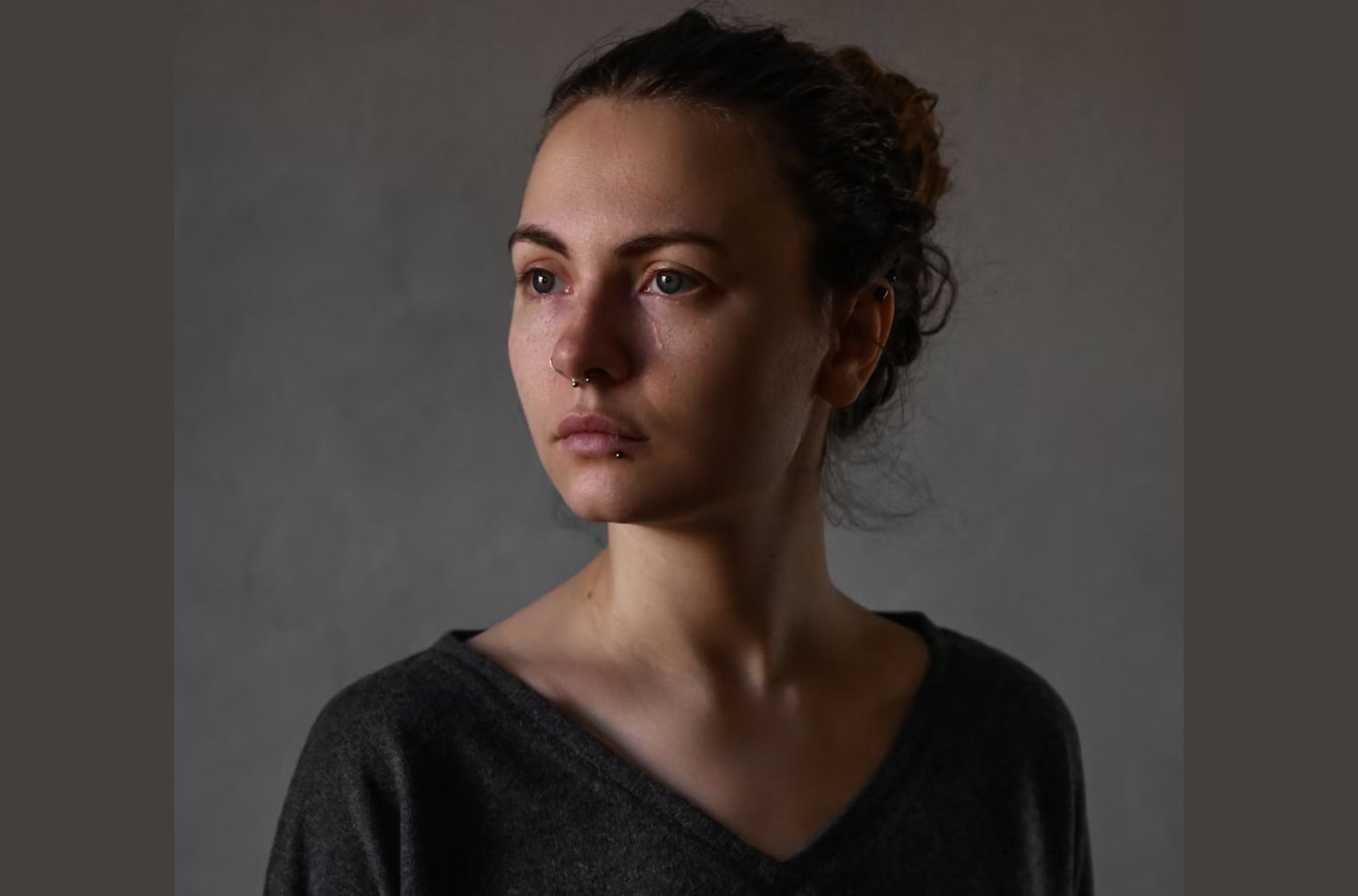 Γυναίκα με σκουλαρίκια στο πρόσωπο και γκρι μπλούζα με άνοιγμα στο λαιμό είναι δακρυσμένη