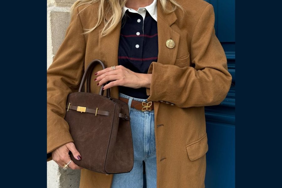 Γυναίκα με καφέ παλτό και τζιν παντελόνι κρατάει καφέ τσάντα με χρυσές λεπτομέρειες