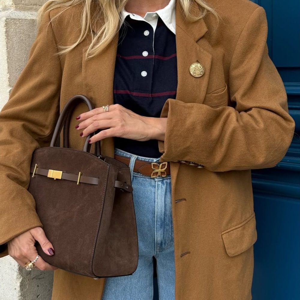 Γυναίκα με καφέ παλτό και τζιν παντελόνι κρατάει καφέ τσάντα με χρυσές λεπτομέρειες