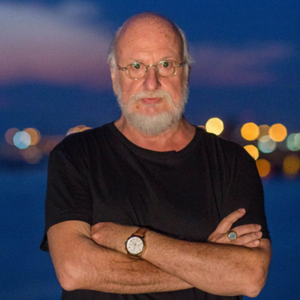 Άντρας με λευκά γένια και σταυρωμένα χέρια, φοράει μαύρο κοντομάνικο μπλουζάκι, γυαλιά μυωπίας και ρολόι στο χέρι