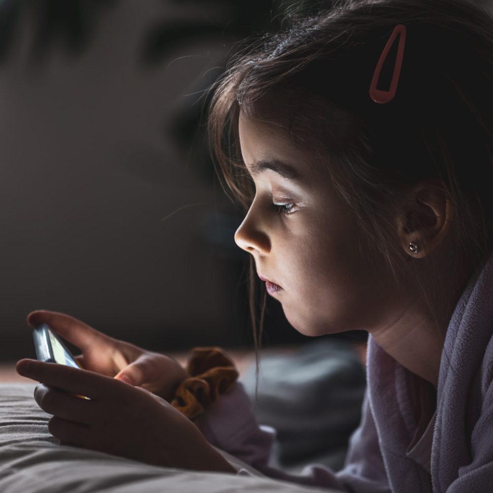 Little girl uses smartphone, lying on pillow at home, gadgets addiction concept.