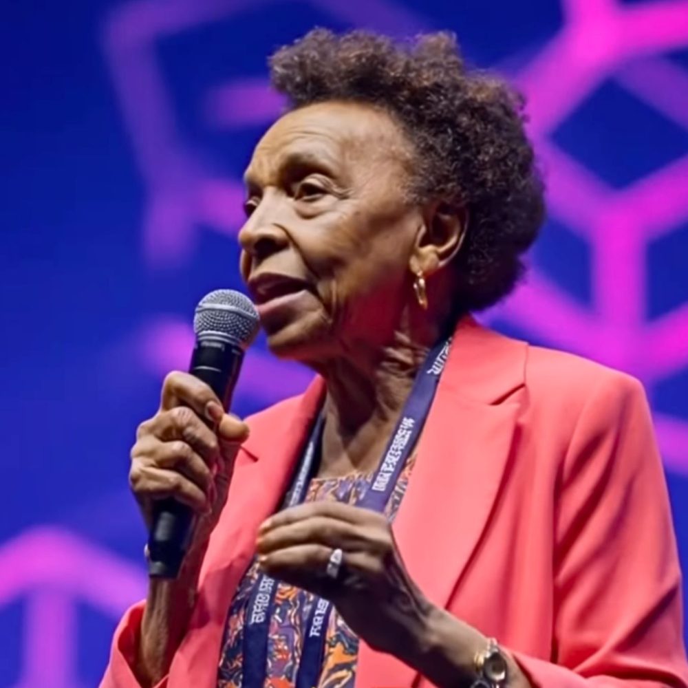 Γυναίκα με κοραλί σακάκι μιλάει κρατώντας μικρόφωνο στο χέρι της