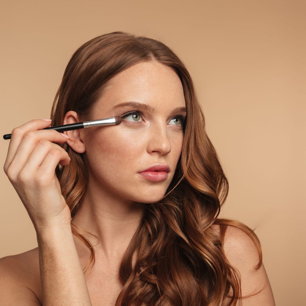 Beauty portrait of mystery ginger woman with long hair looking away while applying cosmetics with brush over cream background
