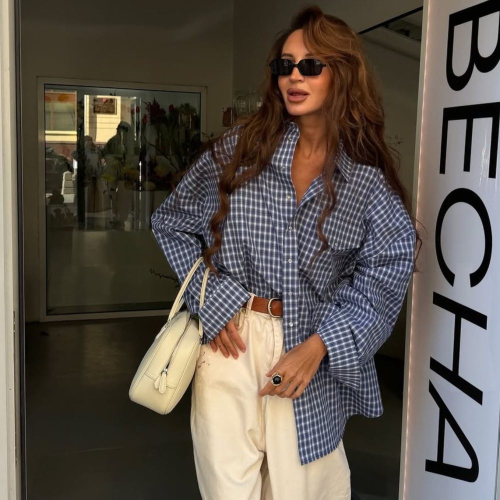 Γυναίκα με καστανά μαλλιά φοράει καρό πουκάμισο, λευκό παντελόνι, λευκή τσάντα και μαύρα γυαλιά ηλίου