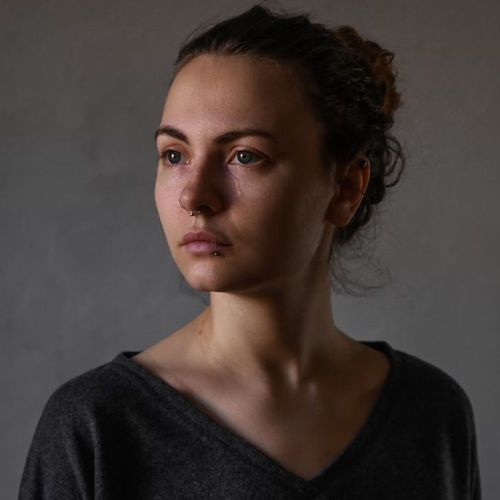 Γυναίκα με σκουλαρίκια στο πρόσωπο και γκρι μπλούζα με άνοιγμα στο λαιμό είναι δακρυσμένη